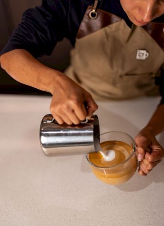 A barista pours steamed milk into a cup of coffee, creating latte art with a small pitcher overhead.