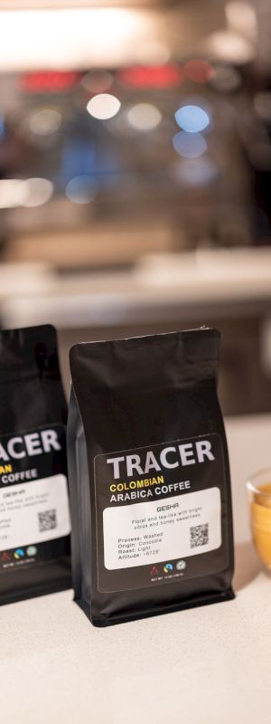 Two bags of Tracer coffee (Colombian Arabica) sit on a counter beside a glass of espresso, with a blurred cafe backdrop in the background.