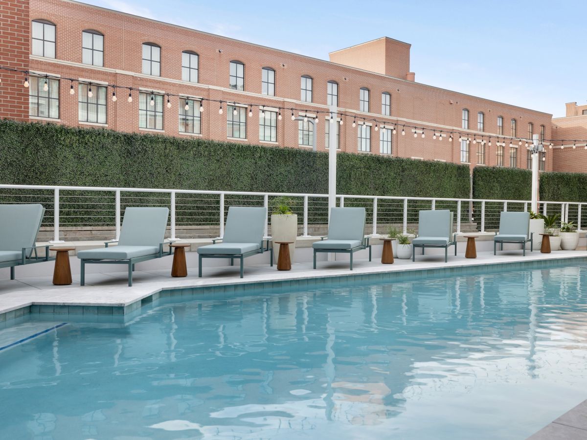 A resort-style pool area with blue water, multiple lounge chairs, and umbrellas along the deck, set against a brick building backdrop.