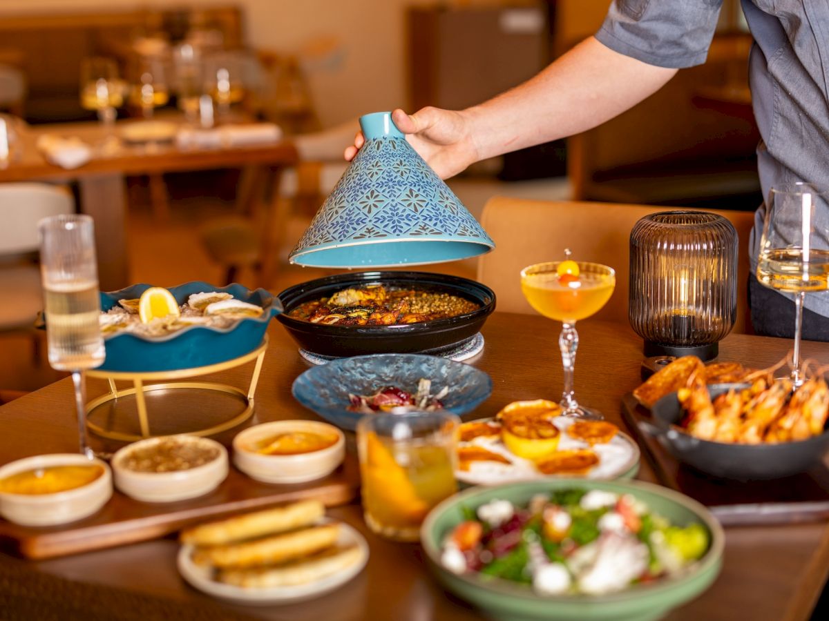 A stylish spread with tapas and bowls, a pot and a blue cloche being lifted, plus cocktails, bread, dips, and salads on a cozy dining table.