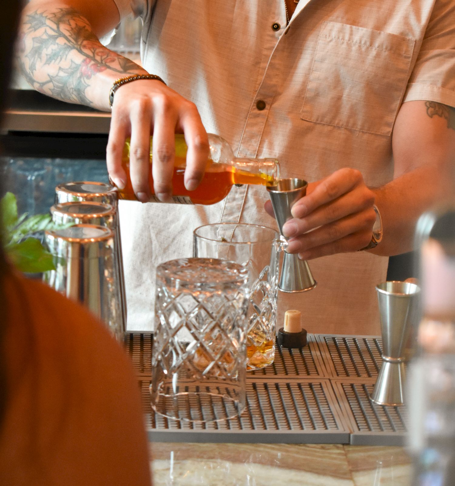 A bartender pours a bright orange drink into a glass behind a marble bar, with bottles and bar tools visible in the background.