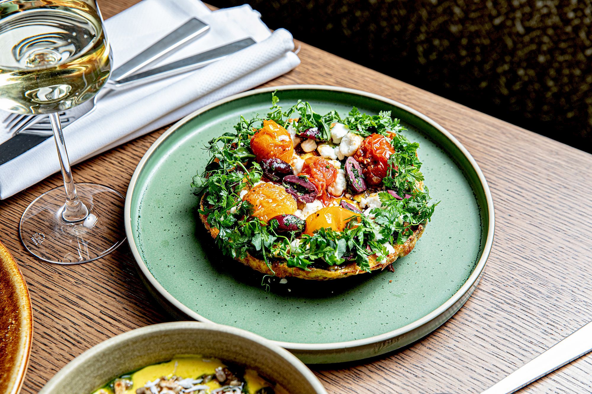 A veggie-filled dish on a green plate with greens, roasted veggies, and a sunny yolk centerpiece, beside a glass of white wine on a wooden table.