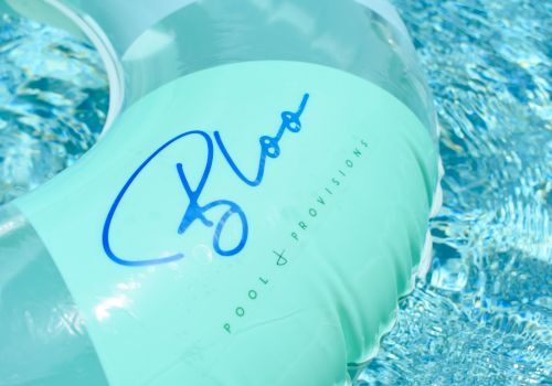 A light blue pool float with blue handwritten-style logo and the words &ldquo;ROOM + BROPTISION&rdquo; on it, resting on a shimmering pool surface.