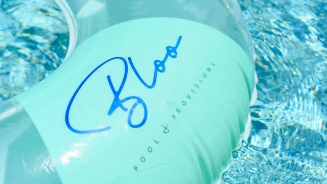 A light blue pool float with blue handwritten-style logo and the words &ldquo;ROOM + BROPTISION&rdquo; on it, resting on a shimmering pool surface.