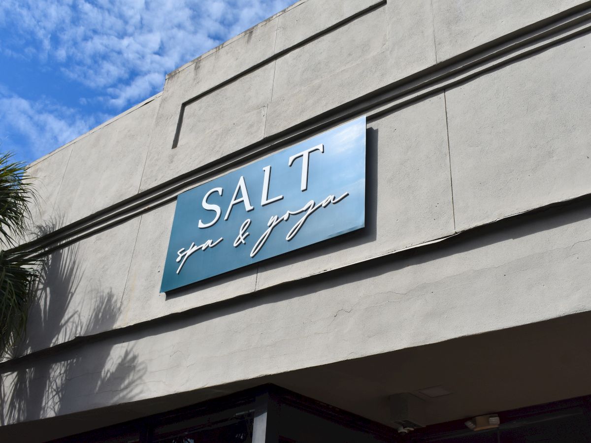 A storefront with a blue sign reading &ldquo;SALT&rdquo; and cursive text beneath, against a concrete building with a clear blue sky.