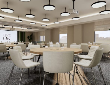 A modern conference room with round tables, cream chairs, projector screen, and large windows; plants add a touch of greenery, lighting fixtures hang above.