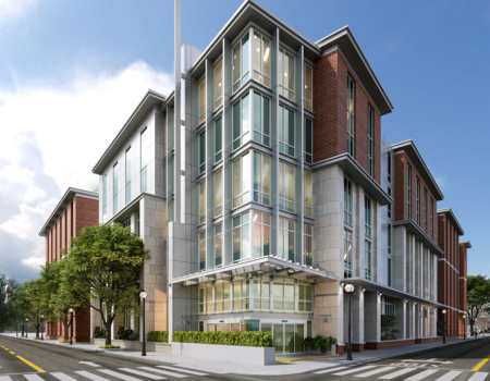 Modern multi-story corner building with glass windows, brick accents, and a small green hedge at the base, on a sunny urban street.
