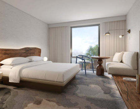 A bright hotel room with a king bed, wooden headboard, night lamps, a seating area, desk, and a large window with sheer curtains.