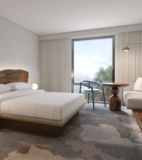 A bright hotel room with a king bed, wooden headboard, night lamps, a seating area, desk, and a large window with sheer curtains.