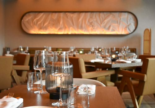 A stylish restaurant dining room with wooden tables, elegant glassware, soft lighting, and upholstered chairs, ready for guests.