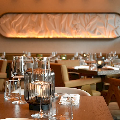A stylish restaurant dining room with wooden tables, elegant glassware, soft lighting, and upholstered chairs, ready for guests.