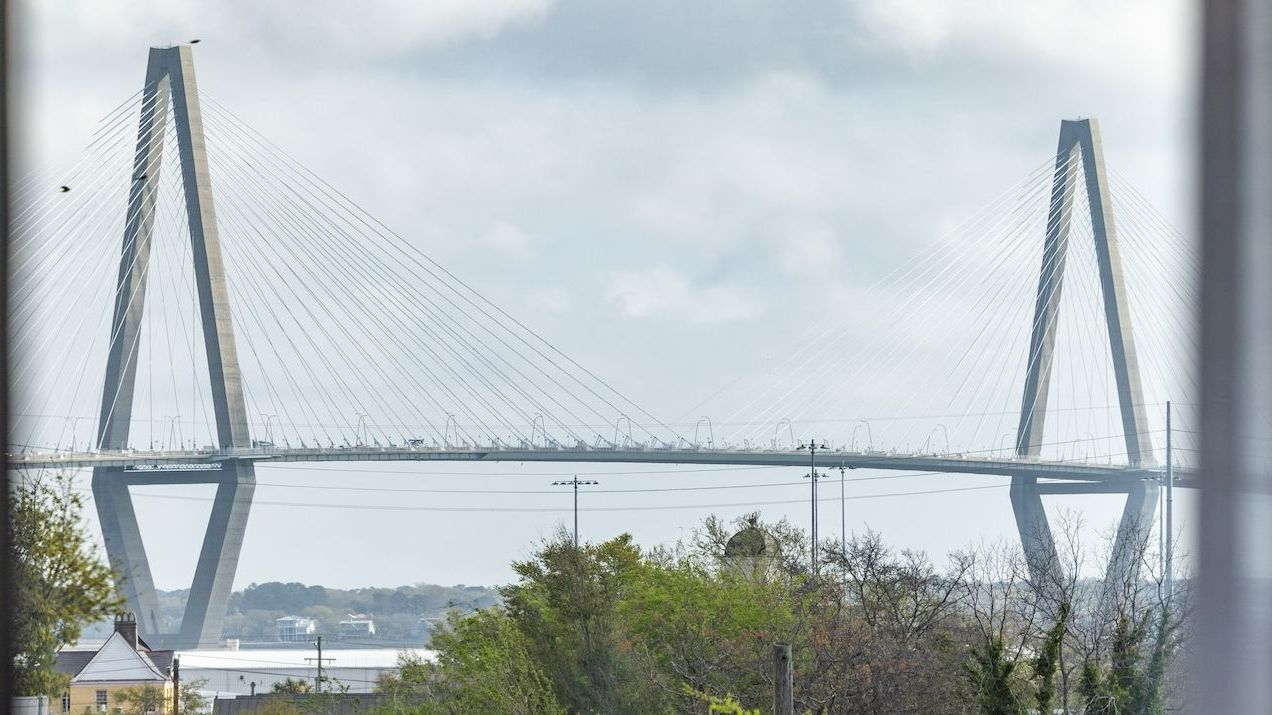 A cable-stayed suspension bridge with tall towers rises over the river, framed by trees under a cloudy sky.