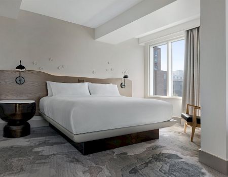 A modern hotel bedroom with a large bed, two wall lamps, a small chair, and a large window with curtains.
