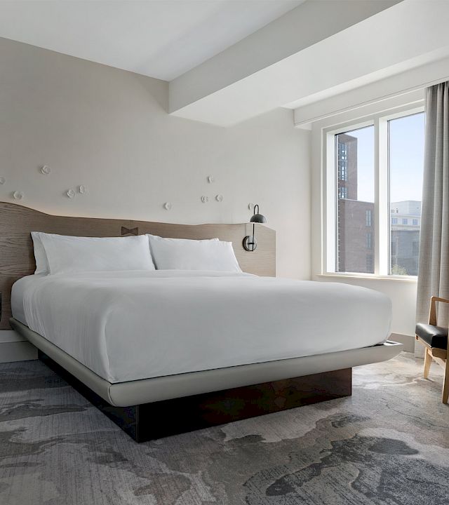A modern hotel bedroom with a large bed, two wall lamps, a small chair, and a large window with curtains.