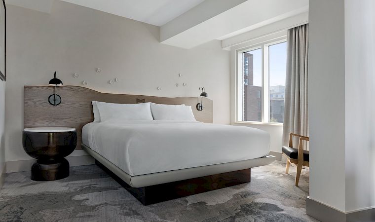 A modern hotel bedroom with a large bed, two wall lamps, a small chair, and a large window with curtains.