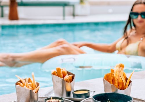 A woman relaxes in a pool while a table in front holds fried snacks, fries, chicken wings, and dipping sauces.