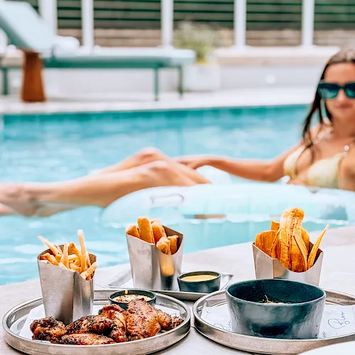 A woman relaxes in a pool while a table in front holds fried snacks, fries, chicken wings, and dipping sauces.