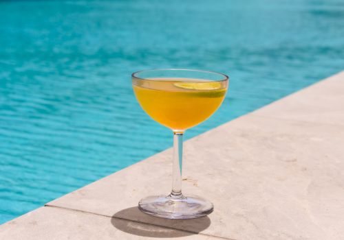 A glass of orange cocktail with a lime garnish sits poolside on a concrete edge.