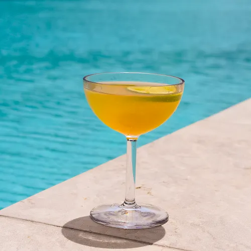 A glass of orange cocktail with a lime garnish sits poolside on a concrete edge.