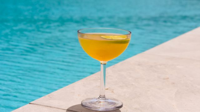 A glass of orange cocktail with a lime garnish sits poolside on a concrete edge.