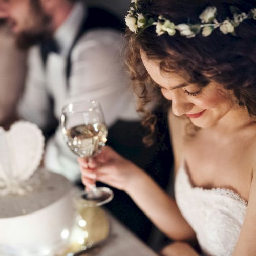 A bride in a floral crown smiles while holding a champagne glass at a candlelit reception.