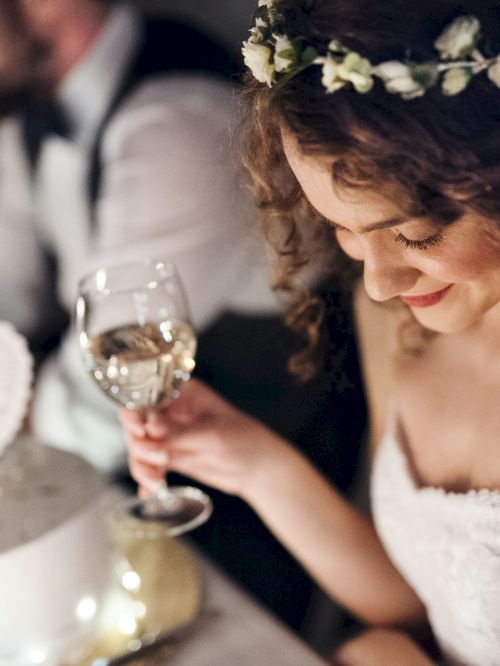 A bride in a floral crown smiles while holding a champagne glass at a candlelit reception.