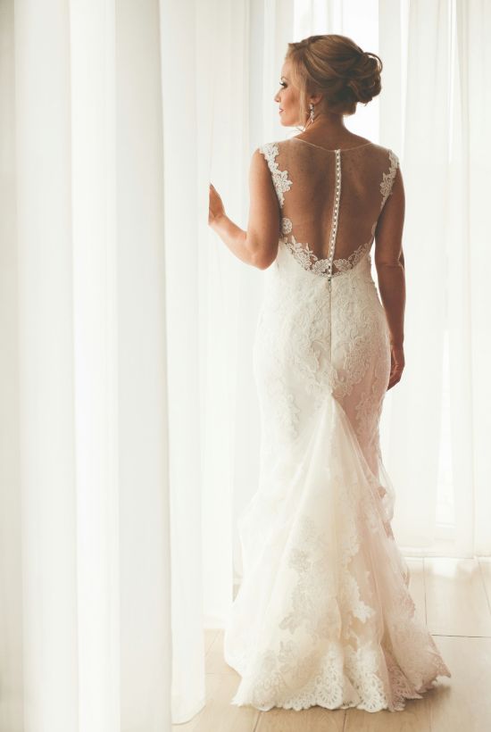 A woman in a lace back wedding dress stands by bright curtains, seen from behind.