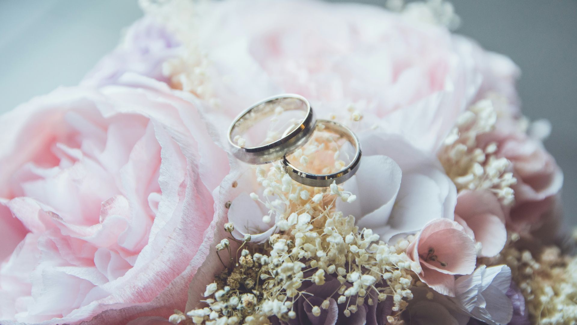 Two wedding rings sit on a floral arrangement, held in hands surrounded by pastel pink blossoms and delicate white flowers.