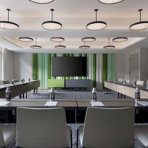 A modern conference room with large tables, gray chairs, pendant lights, a big TV screen, and green-and-gray wall accents.