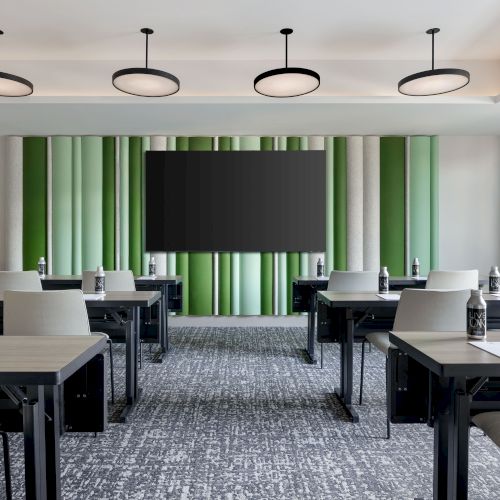 A modern conference room with caf&eacute; tables and chairs, green-striped curtains, a large TV screen, blue-gray carpet, and four ceiling lights.