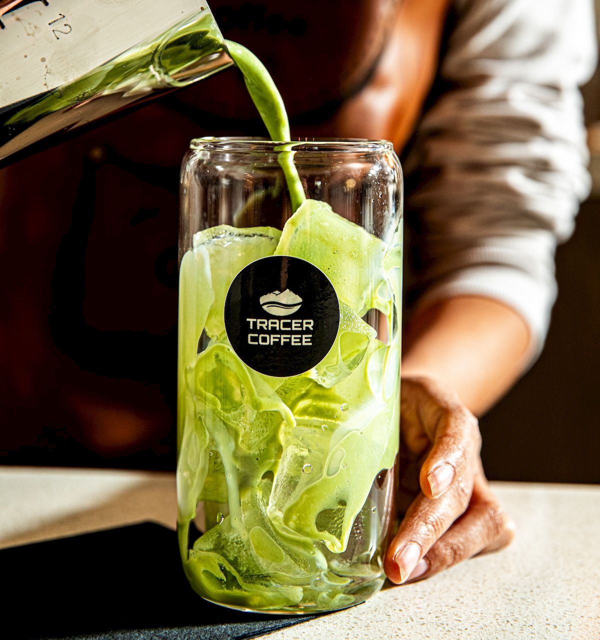 A person pours green liquid into a bottle labeled &ldquo;Tracer Coffee,&rdquo; with lime slices beside it.