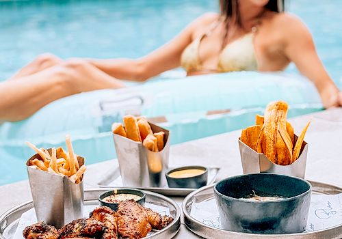 A woman in a pool wearing sunglasses, surrounded by a poolside food spread&mdash;fries, wings, sauces, and snacks.