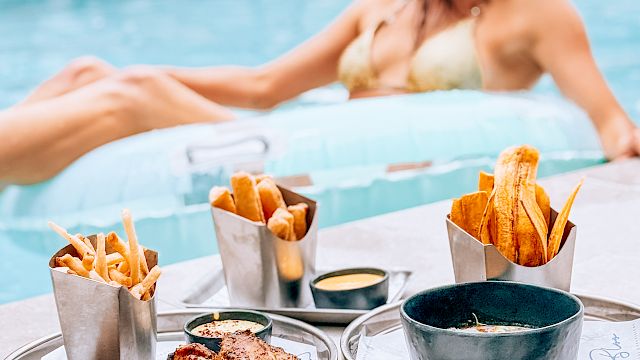 A woman in a pool wearing sunglasses, surrounded by a poolside food spread&mdash;fries, wings, sauces, and snacks.