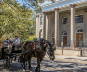 A horse-drawn carriage with passengers rolls past a neoclassical church façade in bright sunshine.