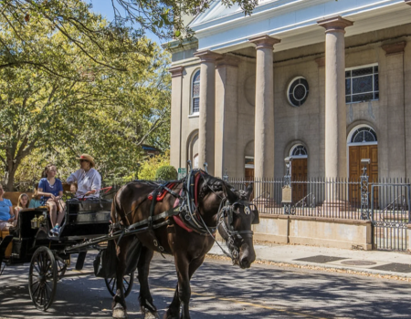 A horse-drawn carriage with passengers rolls past a neoclassical church façade in bright sunshine.