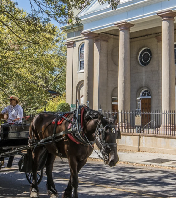 A horse-drawn carriage with passengers rolls past a neoclassical church façade in bright sunshine.