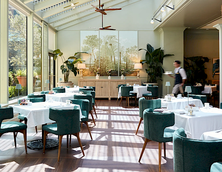 A bright event dining room with set tables, teal chairs, warm lamps, leafy plants, and a mural backdrop under ceiling fans.