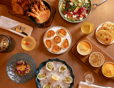 A wooden table spread with assorted meals—salads, flatbreads, fried shrimp, dips, and drinks—served family-style.